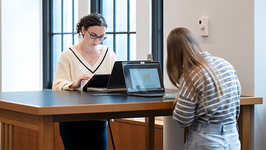 Two researchers work on their laptops across from one another at a standing desk in the library.